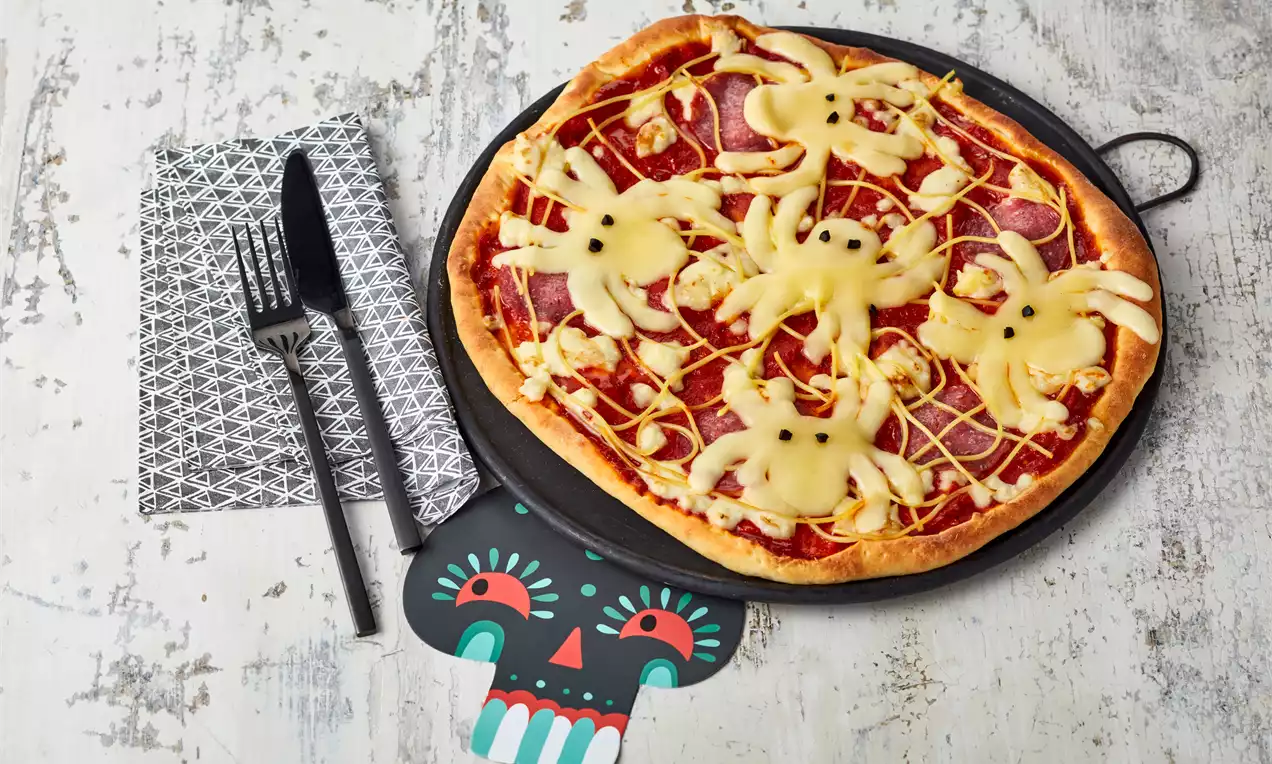 Runde Pizza mit knusprigem Rand und roter Tomatensoße, belegt mit gelben Käse-Spinnen auf schwarzer Platte neben Besteck und Totenkopf-Deko
