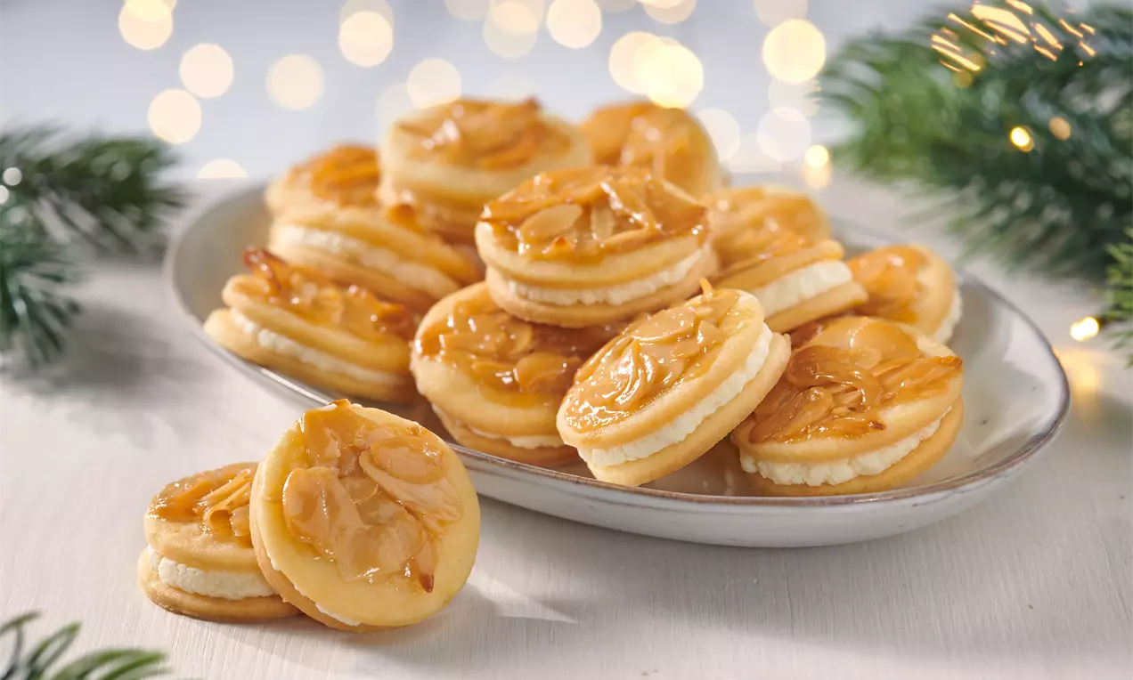 Runde Bienenstich-Plätzchen mit goldbrauner Mandelschicht, heller Füllung und feinem Glanz, arrangiert auf weißer Platte mit grünen Tannenzweigen