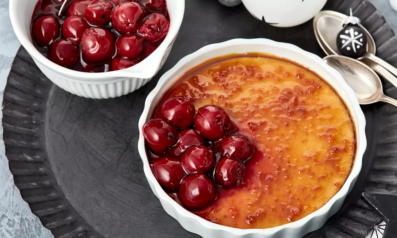 Zimt-Crème-Brûlée mit karamellisierter Oberfläche in weißer Schale, darauf glänzende rote Glühweinkirschen auf dunklem Teller drapiert