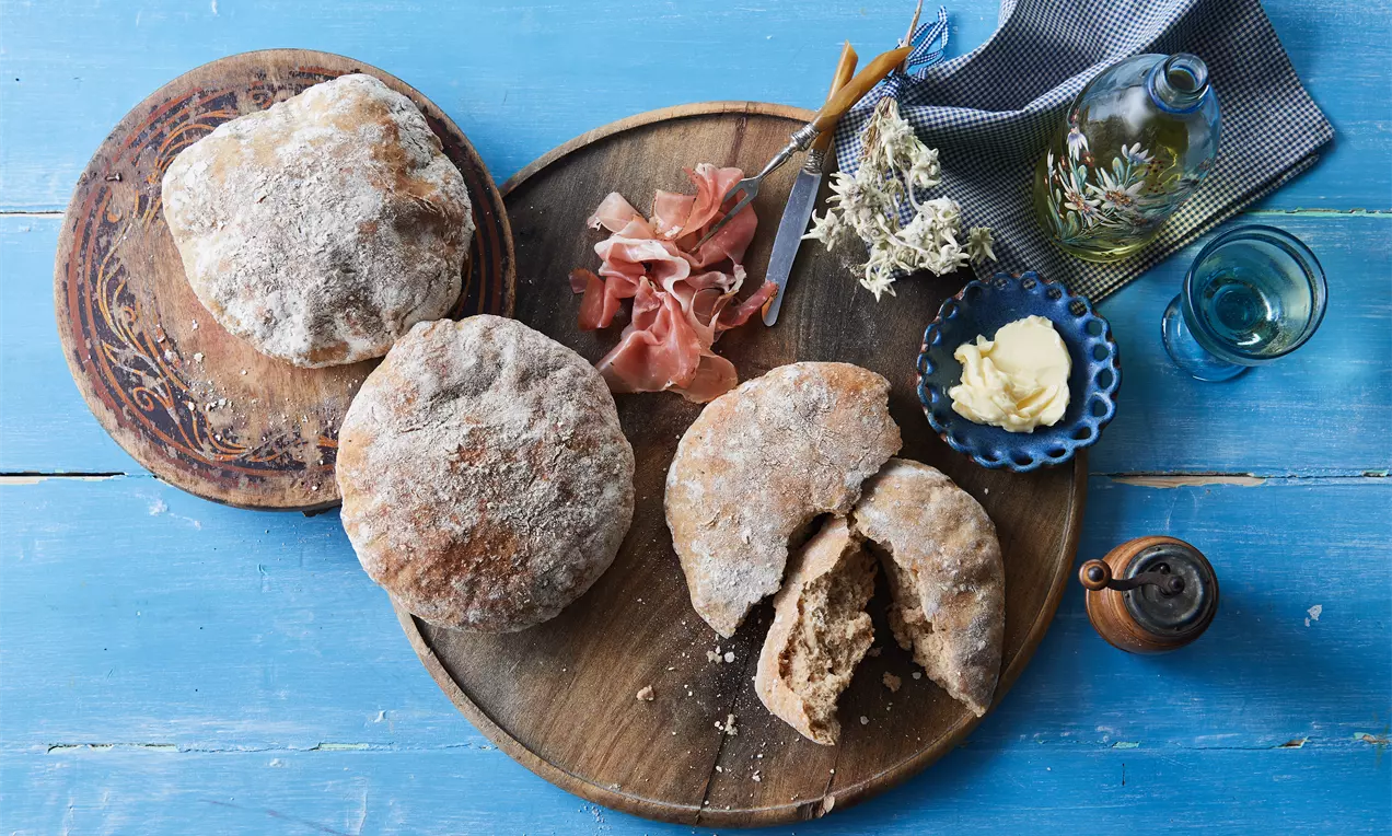 Runde, leicht flache Brote mit bemehlter, goldbrauner Kruste auf rustikalen Holzbrettchen, begleitet von Schinken, Butter und dekorativer Kräuter